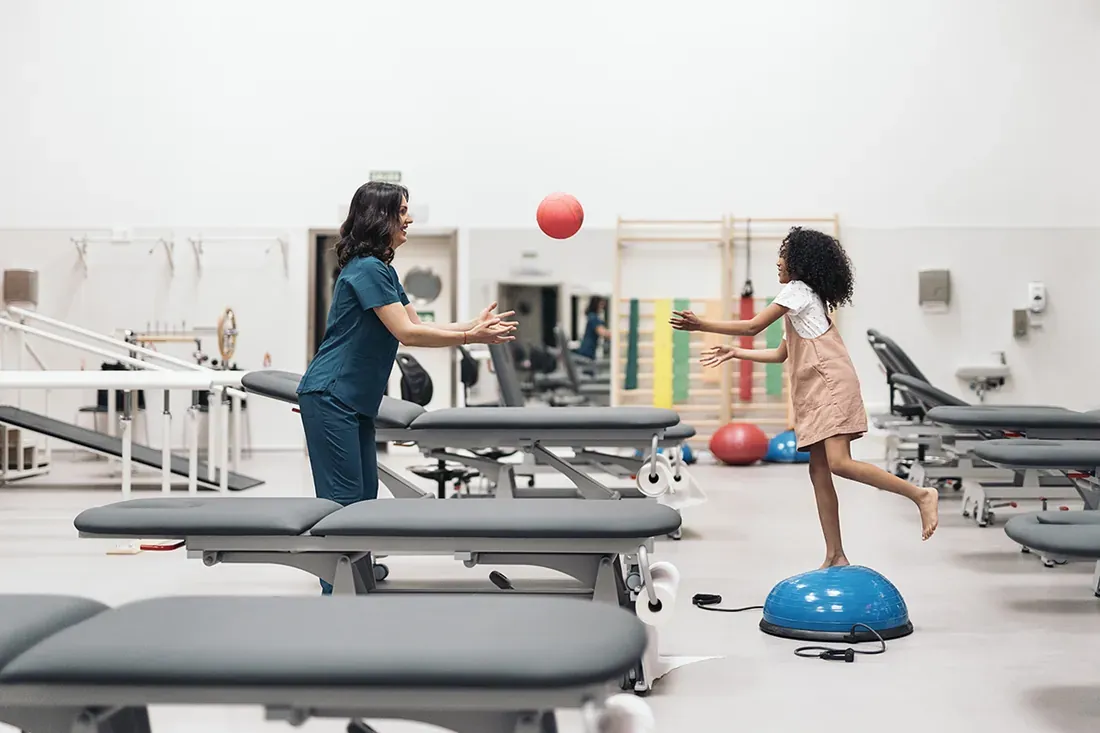 Fisioterapeuta lanza una pelota a una niña en equilibrio en la sala de rehabilitación